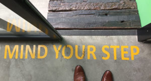 A person observes a "Mind Your Step" sign before walking down the stairs